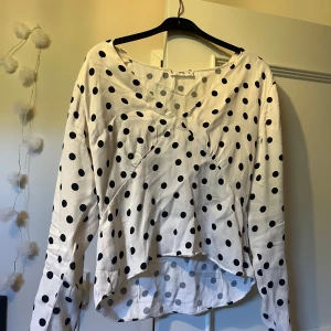 Prickig blus från MNG - Vit blus från MNG med svarta prickar och v-ringning. Blusen har långa ärmar och en lätt figursydd passform med snyggt omlottdetalj framtill. Perfekt för dig som gillar en lekfull men stilren look. Storlek L men är mer som en S eftersom att plagget är litet i modellen