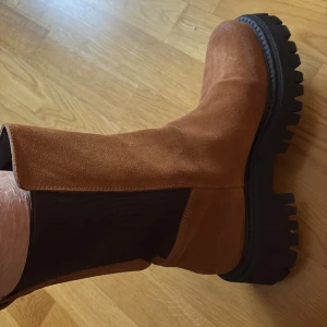 Bruna mocka chelsea boots med grov sula - Säljer ett par snygga chelsea boots i brun mocka med svarta elastiska paneler på sidorna och kraftig, svart räfflad sula. Skorna har rund tå och platt sula, perfekt för dig som vill ha en cool och robust look till hösten eller vintern.Andvänt högst 2 gånger. Köpta i Milano.