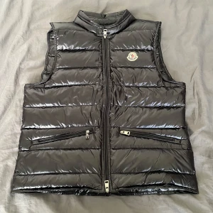 Svart dunväst från Moncler - Snygg svart dunväst från Moncler med glansig finish och klassisk logga på bröstet. Västen har två dragkedjefickor framtill och stängs med dragkedja. Perfekt för lager-på-lager och ger en clean streetstyle-vibe.