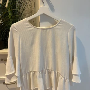 Vit volangblus från Gina Tricot XS - Supersöt vit blus från Gina Tricot i storlek XS. Blusen har volangdetaljer både vid ärmslut och nederkant samt trekvartsärmar. Den är tillverkad i ett mjukt, strukturerat tyg och har en rund halsringning. Perfekt för dig som gillar en feminin och trendig stil.