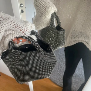 Glittrig svart handväska med kedja - Säljer en liten svart handväska med glittriga stenar och silverfärgad kedja. Väskan har två handtag och ett rymligt fack med svart innerfoder. Perfekt för att lyfta vilken outfit som helst med lite extra bling💖💖💖.