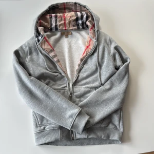 Burberry, Grå hoodie från Burberry med fleece - Snygg grå hoodie från Burberry med dragkedja och klassiskt rutigt foder i huvan. Insidan är fodrad med mjuk vit fleece för extra komfort. Perfekt för dig som vill ha en stilren och lyxig look med ikoniska Burberry-detaljer. Stl L, sitter som M.