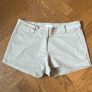 Beige shorts från Nelly i storlek 36 - Snygga beige shorts från Nelly med klassisk design, bälteshällor, framfickor och fejk bakfickor. Tillverkade i bomullsmix och perfekta för varma dagar och enkel att matcha med allt.