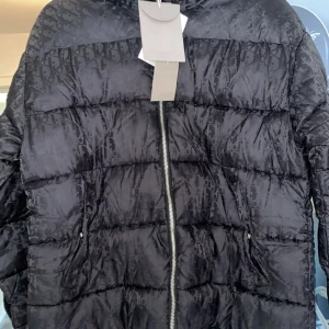 Svart pufferjacka från Dior - Exklusiv svart pufferjacka från Dior med diskret monogrammönster över hela jackan. Jackan har dragkedja framtill med Dior-logga, två fickor och patch med Dior på ärmen. Perfekt för dig som vill ha en lyxig och trendig vinterjacka.
