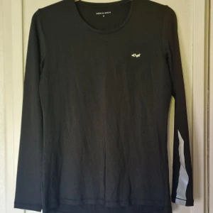 Svart långärmad träningströja Röhnisch - Svart långärmad träningströja från Röhnisch med rund halsringning och logga på bröstet. Reflexdetaljer på ärmarna ger en sportig vibe. Tillverkad i mjukt och stretchigt syntetmaterial som andas, perfekt för aktiva dagar. Endast använd ett fåtal gånger.