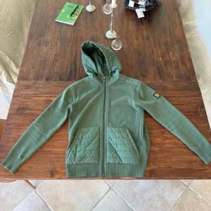 Grön hybridjacka/Cardigan från Belstaff Strl S, Passar M - Snygg grön hybridjacka från Belstaff med quiltade detaljer på huva och fickor. Jackan har dragkedja framtill, ribbade muddar och lång ärm. Materialmix av stickat och syntet ger en sportig och modern look. Perfekt för lager-på-lager under kyliga dagar.
