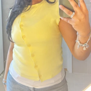 Gul ärmlös topp med sned knäppning - Snygg gul ärmlös topp med sned knäppning framtill och matchande knappar. Mjuk och stretchig, perfekt till jeans eller kjol. Rund halsringning och croppad passform ger en fräsch och trendig look.