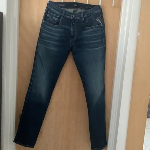 Replay Hyperflex Anbass jeans blå - Säljer ett par Replay Hyperflex Anbass jeans i mörkblå tvätt med snygga slitningar. Klassisk femficksmodell med smal passform och stretchigt material för extra komfort. Märkesdetaljer på bakficka och midjeband. Perfekta för dig som gillar stilrena jeans med modern känsla.