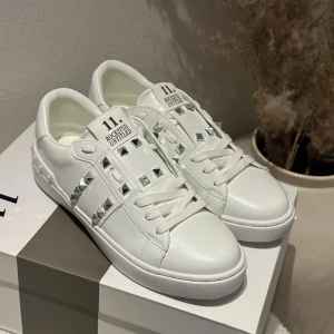 Valentino Rockstud vita sneakers - Snygga vita Valentino Garavani Rockstud sneakers i skinn med ikoniska silverfärgade nitar och platt sula. Klassisk rund tå, snörning och coola detaljer längs sidor och häl. Perfekta för dig som vill sticka ut med en lyxig touch.