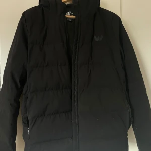 Svart pufferjacka från Whistler - Svart pufferjacka från Whistler med huva och dragkedja framtill. Jackan har en stilren design med diskret logga på bröstet och två fickor med dragkedja. Perfekt för kalla dagar när du vill hålla värmen och ändå se snygg ut.