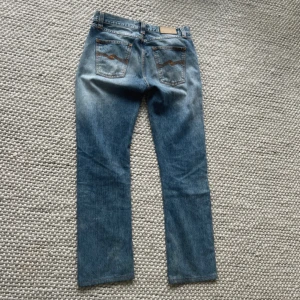 Blå jeans från Nudie Jeans straight fit - Säljer ett par klassiska blå jeans från Nudie Jeans med raka ben och snyggt slitna detaljer. Jeansen har fem fickor, orange sömmar och Nudie-logga på bakfickorna. Tillverkade i bomull med en skön passform och normal midja.
