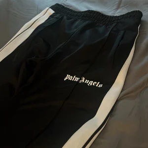 Palm Angels svarta trackpants med vita ränder - Svarta trackpants från Palm Angels med vita sidoränder och logga broderad på benet. Byxorna har elastisk midja och raka ben, tillverkade i ett syntetmaterial som ger en sportig vibe. Perfekta för en streetwear-look.pris ej hugget !