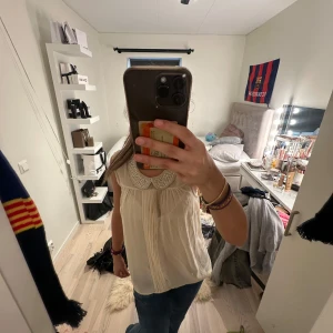 Beige blus med pärlkrage och knyt - Supersöt ärmlös beige blus med glittrande pärlkrage och fina veck framtill. Blusen har knyt i nacken och är gjord i ett tunt, flowy material som känns lyxigt. Perfekt för dig som gillar detaljer och vill ha något extra till outfiten.