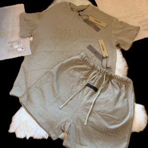 essentials fear of god shorts och t-shirt - Snygga beige shorts och t shirt från Fear of God med broderad logga framtill. Modellen har elastisk midja med snörning och är tillverkad i mjuk bomull för skön komfort. Perfekt för chill dagar och streetwear-stil.