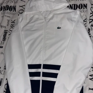 Vit vindjacka från Lacoste Sport - Lacoste Tracksuit. Snygg vit svart vindjacka och byxor från Lacoste med luva och dragkedja. Jackan har svarta detaljer och stora Lacoste-trycket på ryggen. Klassisk krokodillogga på bröstet. Tillverkad i polyester och perfekt för dig som gillar sportig och fet stil.