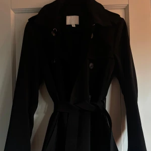 Svart trenchcoat från H&M, strl 34 - Svart trenchcoat från H&M med klassisk krage, dubbelknäppning och bälte i midjan med snyggt spänne. Långärmad modell i slät polyester, perfekt för höst och vår. Stilren och tidlös design som lyfter varje outfit.