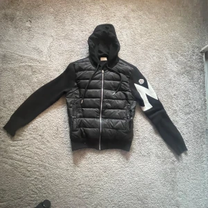 Moncler cardigan - Svart pufferjacka från Moncler med stickade ärmar och huva. Jackan har dragkedja framtill, quiltad front och en vit Moncler-logga på ena ärmen. Snygg kontrast med vit detalj på ärmen och dragsko i huvan. Perfekt för dig som vill ha en stilren och sportig look.