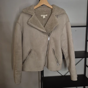 Beige teddyjacka med dragkedja från H&M - Säljer en beige teddyjacka från H&M med mjukt foder och krage i teddy. Jackan har sned dragkedja framtill och två fickor med dragkedja. Perfekt för kyliga dagar och har en clean, stilren look.