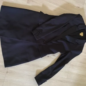 Svart kavajklänning med knappar - Stilren svart kavajklänning med markerad midja och klassisk krage. Långa ärmar och knappar vid ärmslut samt framtill. Perfekt för dig som gillar en dressad look med snygg siluett.