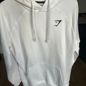 Vit hoodie från Gymshark - Säljer en stilren vit hoodie från Gymshark. Den har en klassisk design med en känguruficka framtill och justerbar huva med dragsko. Perfekt för en avslappnad look eller träning.