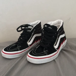 Vans Sk8-Hi svart/vit med röd blixt - Säljer ett par Vans Sk8-Hi sneakers i svart mocka och canvas med vit sula och cool röd blixtdetalj på sidan. Klassisk vit sidestripe, svarta skosnören och kontrastsömmar. Perfekta för dig som gillar streetstyle och vill sticka ut lite extra.