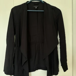 Svart cardigan jacka från Amisu - Svart långärmad cardigan jacka från Amisu med öppen front och snyggt fall. Jackan har dragkedjedetaljer i sidorna och en avslappnad passform. Perfekt för lager-på-lager och enkel att matcha med olika outfits.