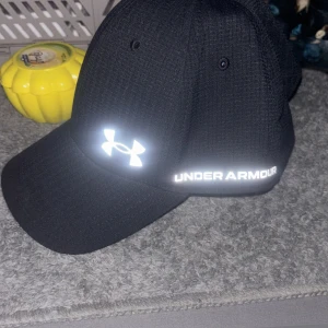 Svart keps från Under Armour - Svart keps från Under Armour med böjd skärm och reflekterande logga samt text framtill. Kepsen är tillverkad i ett ventilerande syntetmaterial med diskret rutstruktur, perfekt för träning eller sportiga outfits.