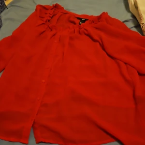 Röd blus med volang från H&M - Snygg röd blus från H&M i storlek 34. Blusen har volangdetaljer vid halsringningen, knappar framtill och är tillverkad i ett lätt och luftigt tyg. Perfekt för dig som vill sticka ut med färg och stil.