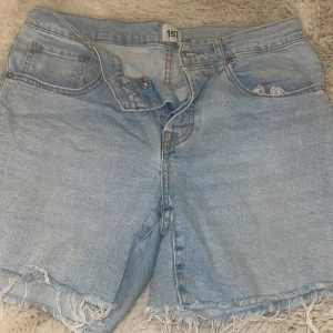 Ljusblå jeansshorts från 157 - Säljer ett par ljusblå jeansshorts från 157 med råa, fransiga benslut och klassisk femficksdesign. Shortsen har knappgylf och en avslappnad passform, perfekta för varma dagar. Tidlös denimstil som är enkel att matcha.
