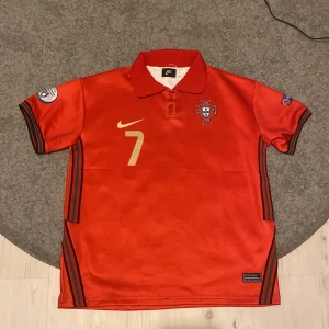Ronaldo portugal tröja - Snygg röd Portugal fotbollströja från Nike med nummer 7 och Ronaldo på ryggen. Tröjan har korta ärmar, krage och svarta detaljer längs sidorna och ärmsluten. Officiella märken på ärmarna och broderad landslagssköld på bröstet.