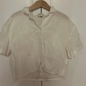 Vit kortärmad skjorta från H&M - Vit skjorta från H&M med korta ärmar och klassisk krage. Skjortan har knäppning framtill och en snygg knytning nedtill för en chill och trendig look. Perfekt till sommaren och enkel att styla med jeans eller kjol.