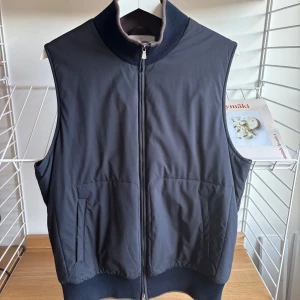 Gran Sasso Väst - Stilren mörkblå dunväst från Gran Sasso med brunt stickat foder och hög krage. Västen har två fickor framtill. Perfekt för lager-på-lager och snygg till både hoodie och skjorta. Köpt på Ströms så kvitto kan nog letas fram.