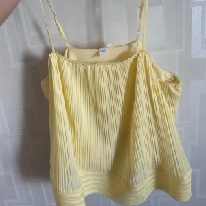 Gul plisserad linnetopp från YAS - Superfin ljusgul linnetopp från YAS med tunna, justerbara axelband och plisserat tyg. Toppen har en luftig och flowy passform, perfekt till sommaren. Snygg att styla med jeans eller kjol för en fräsch look. Aldrig använd och kostar 600kr nypris