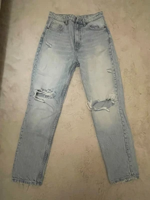 Ljusblå ripped jeans med raka ben - Säljer ett par ljusblå jeans med hög midja och raka ben. Jeansen har slitna detaljer och stora hål vid knäna för en trendig, avslappnad look. Klassisk femficksmodell och tillverkade i bomullsdenim. Perfekta för dig som gillar streetstyle.