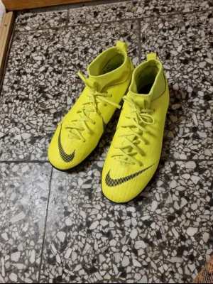 Nike Mercurial gula fotbollsskor - Säljer ett par Nike Mercurial fotbollsskor i en riktigt skarp gul färg med svarta detaljer och logga. Skorna har snörning, platt sula och en strumpliknande högre krage för extra stöd. Tillverkade i syntetmaterial som är lätt och smidigt för snabba rörelser på planen. De är I storlek 38,5. Det står namn inuti skorna.