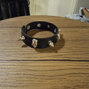Svart skinnarmband med nitar och döskallar - Säljer ett svart armband i PU skinn med silverfärgade nitar och döskalle-detaljer. Armbandet har tryckknappar för justering och passar perfekt för dig som gillar edgy stil. Bredden är rejäl och materialet är syntetiskt skinn. 22 cm lång.