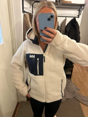 Vit teddyjacka från Helly Hansen - Säljer en fluffig vit teddyjacka från Helly Hansen med marinblå detaljer och bröstficka med dragkedja. Jackan har huva, hel dragkedja framtill och två sidofickor. Perfekt för kalla dagar och riktigt mysig att bära.
