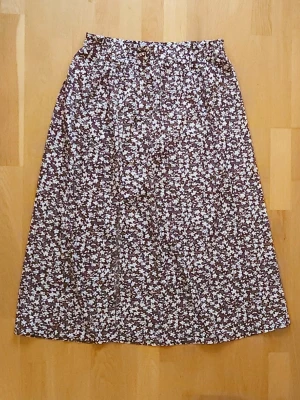 Kjol - En flowy långkjol med slit och brunvitt blommönster. Kjolen har en bekväm resår i midjan och är gjord i ett lätt tyg som ger en skön känsla. Perfekt för dig som gillar en avslappnad men ändå söt stil.