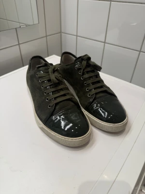 Lanvin sneakers i mörkgrön mocka - Säljer ett par snygga Lanvin sneakers i mörkgrön mocka med lackade tåpartier och vita gummisulor. Skorna har rund tå, snörning och cool kontrast mellan mocka och glansigt material framtill. Perfekta för dig som vill ha något unikt och stilrent.