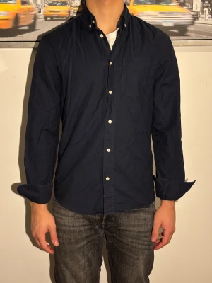 Mörkblå skjorta med knappar - Snygg mörkblå skjorta med klassisk button-down krage och vita knappar. Skjortan har en bröstficka och är långärmad med enkel, rak passform. Tillverkad i mjuk bomull som känns skön mot huden. Perfekt att bära öppen över en t-shirt eller stängd för en clean look.