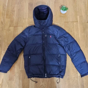 Svart pufferjacka från Polo Ralph Lauren - Säljer en marinblå pufferjacka från Polo Ralph Lauren med huva och dragkedja framtill. Jackan har tre fickor med dragkedja, snörning i nederkant och det klassiska Polo-logot broderat på bröstet. Perfekt för kalla dagar och riktigt snygg streetstil.
