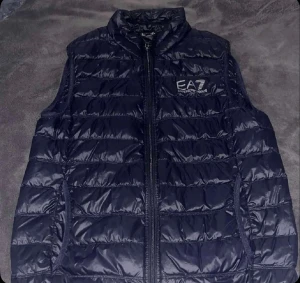 Mörkblå dunväst från EA7 Emporio Armani - Snygg mörkblå dunväst från EA7 Emporio Armani med quiltad design och hög krage. Västen har dragkedja framtill och två sidofickor. Perfekt för att addera lite extra stil till din outfit. Logotypen är broderad på bröstet för en exklusiv touch.