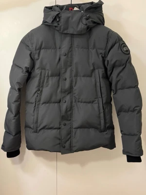 Svart dunjacka från Canada goose - Säljer en svart dunjacka från Canada goose med huva och snygga detaljer som snörning och dragkedja. Jackan har quiltad design, långärmade ärmar och två praktiska fickor med dragkedja. Perfekt för kalla dagar och har en cool patch på ärmen.