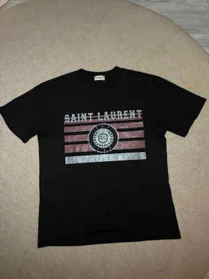 Svart Saint Laurent t-shirt med tryck - Svart t-shirt från Saint Laurent med coolt tryck framtill i vitt och rött, samt texten 'SAINT LAURENT' och emblem. Klassisk passform med rund hals och korta ärmar. Perfekt för dig som vill ha en stilren men edgy look.