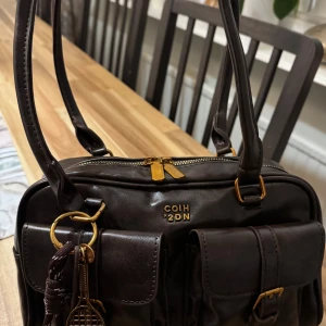 Brun axelväska från COIH 2DN - Snygg brun axelväska i skinn med guldfärgade detaljer och dubbla framfickor med spännen. Väskan har två långa axelremmar och dragkedja upptill. Dekorerad med nyckelring och tennisracket-hänge för extra stilpoäng.
