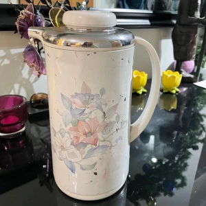 Blommig vit termos från Tiger - Snygg retro termos från Tiger i vit plast med blommigt mönster i pastellfärger. Har ett handtag och lock i vitt och silverfärgad metall upptill. Perfekt för att hålla drycken varm och har en klassisk, stilren form med mjuka linjer.