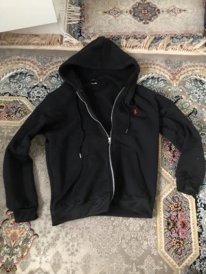 Svart hoodie från Ralph Lauren - Svart hoodie från Ralph Lauren med dragkedja och huva. Klassisk röd logga broderad på bröstet och två fickor framtill. Perfekt för en chill och avslappnad stil.