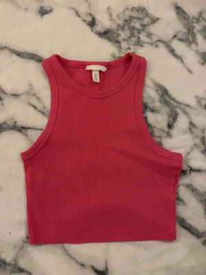 Rosa ribbad croppad linnetopp H&M - Säljer en snygg croppad linnetopp från H&M i en stark rosa färg. Toppen är ribbad och ärmlös med rundad halsringning, perfekt för att matcha med jeans eller kjol. Materialet är mjukt och stretchigt, vilket gör den superbekväm att bära.