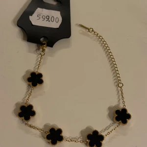 Svart blommigt / klöver armband med guldkedja - Snyggt armband med fem svarta blommor som hänger på en tunn guldfärgad kedja. Blommorna har en mjuk, rundad form och är inramade med små guldfärgade detaljer. Perfekt accessoar för att lyfta din outfit med en cool och elegant touch.