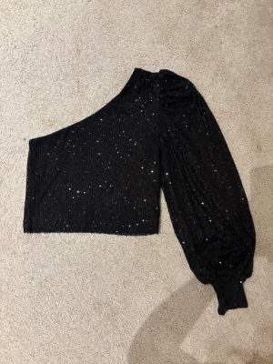 Svart glittrig oneshoulder topp - Säljer en svart oneshoulder topp med glittriga paljetter över hela tyget. Toppen har en lång puffärm med bred mudd och är croppad i modellen. Perfekt för dig som vill ha en fixad topp till new year.